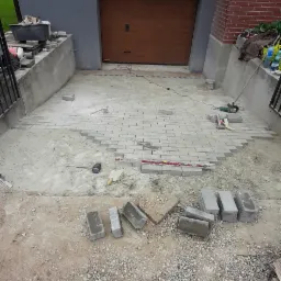 Układanie szarej kostki brukowej na podjeździe przed garażem, widoczne narzędzia i niedokończony fragment nawierzchni.