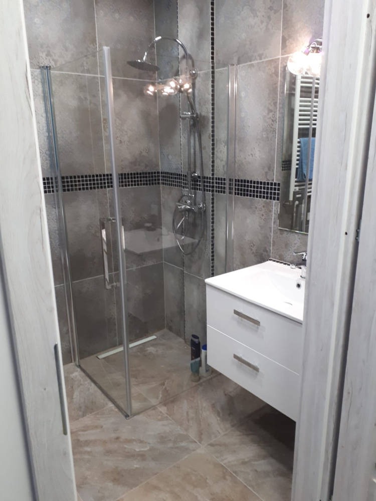 Nowoczesna łazienka z kabiną prysznicową typu walk-in, umywalką z szafką w kolorze białym, szarymi płytkami ściennymi z czarnymi akcentami i beżową podłogą z płytek imitujących kamień.