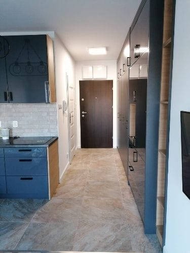 Wnętrze mieszkania po remoncie: widok na przedpokój z kuchnią, szare płytki podłogowe, zabudowa kuchenna w odcieniach granatu, ciemne drzwi wejściowe, szafa w zabudowie w przedpokoju z oświetleniem.