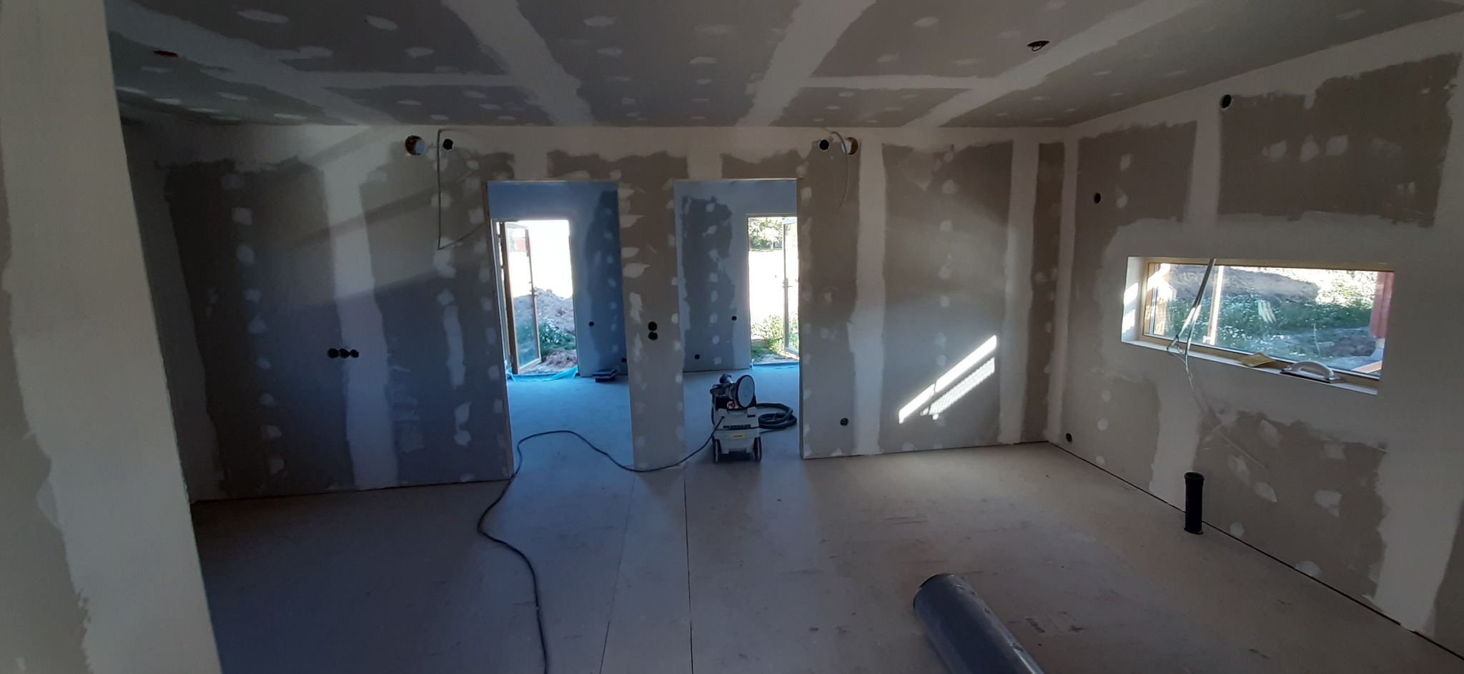 Wnętrze w trakcie prac wykończeniowych: ściany z płyt karton-gips, częściowo wyszpachlowane, widoczne otwory na instalacje, szlifierka do gładzi i rolka folii ochronnej na podłodze.