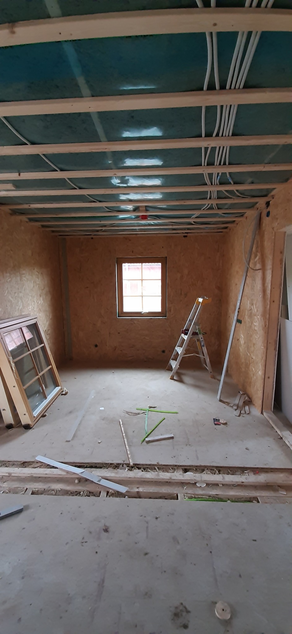 Wnętrze w trakcie remontu: widoczne instalacje elektryczne pod sufitem, okno z widokiem na zewnątrz, drabina, płyty OSB na ścianach i przygotowane ramy okienne.