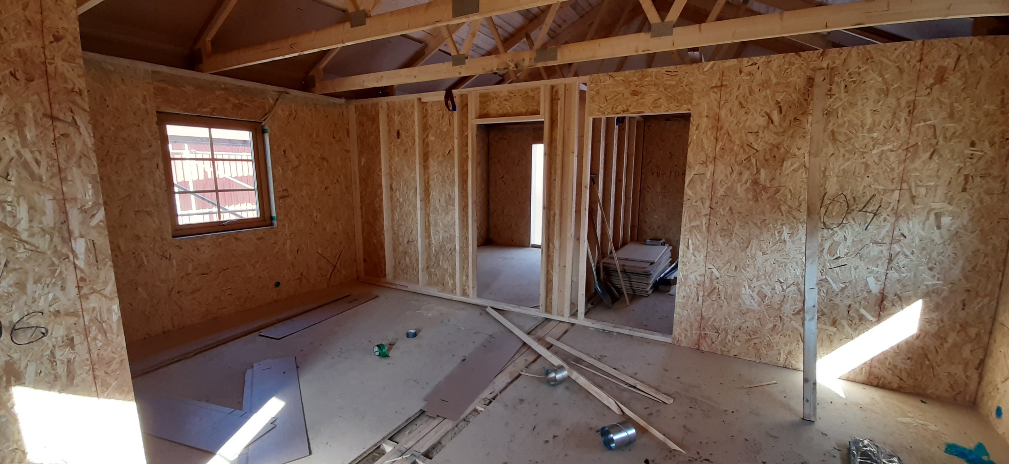 Wnętrze domu w trakcie budowy, ściany wyłożone płytami OSB, widoczne drewniane konstrukcje ścian działowych i stropu, okno z widokiem na zewnątrz, na podłodze resztki materiałów budowlanych.