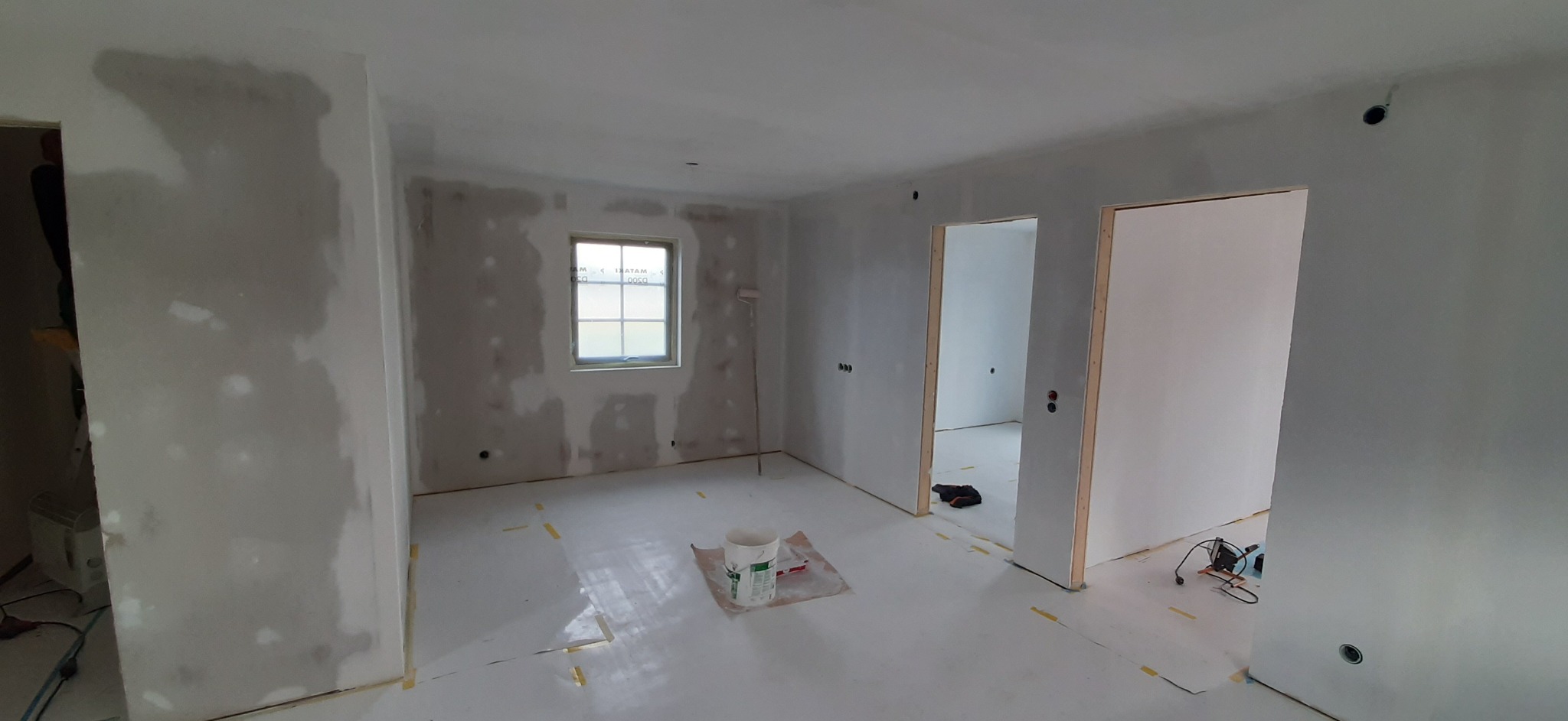 Pokój w trakcie remontu: ściany częściowo pokryte szpachlą, podłoga zabezpieczona folią, widoczny wałek malarski oparty o ścianę, wiadro z farbą na podłodze, narzędzia przy framudze drzwi.