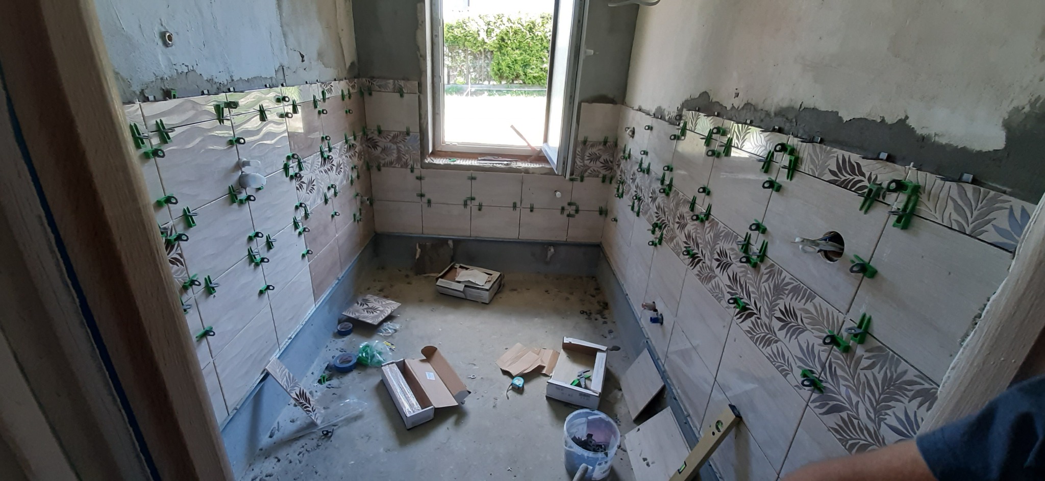 Ściany łazienki w trakcie układania płytek z wzorem liści, widoczne zielone klipsy poziomujące, otwarte pudełka z płytkami na podłodze, okno w tle.