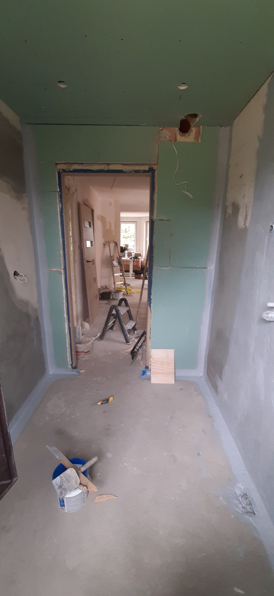 Wnętrze w trakcie prac wykończeniowych: ściany obłożone płytami kartonowo-gipsowymi, widoczne łączenia płyt, drabina, narzędzia malarskie i wiadro z zaprawą, przygotowane do dalszych prac.