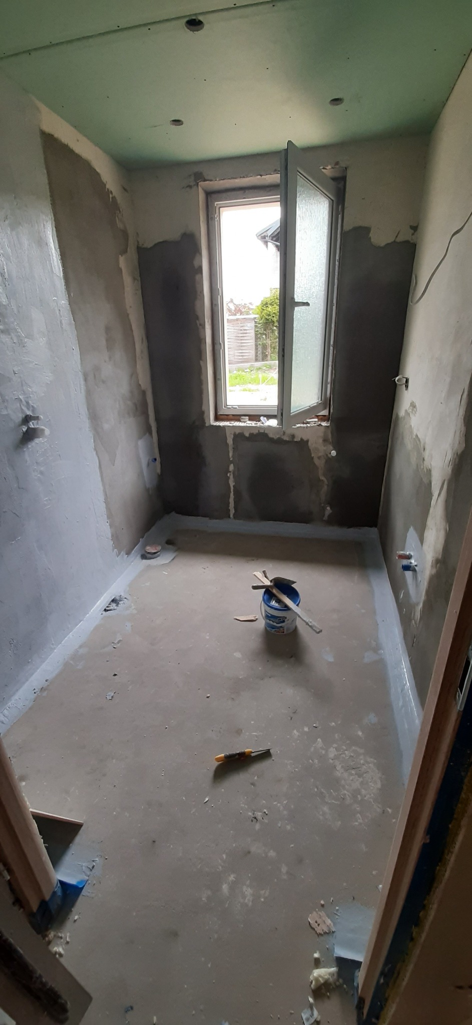 Łazienka w trakcie remontu, widoczne ściany po szpachlowaniu, gruntowanie podłogi, otwarte okno, wiadro z farbą i narzędzia.