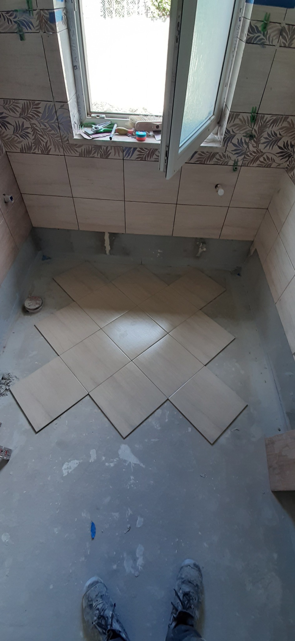 Układanie beżowych płytek podłogowych w łazience, widoczne buty robocze w dolnej części kadru, okno w górnej części kadru, etap prac wykończeniowych.