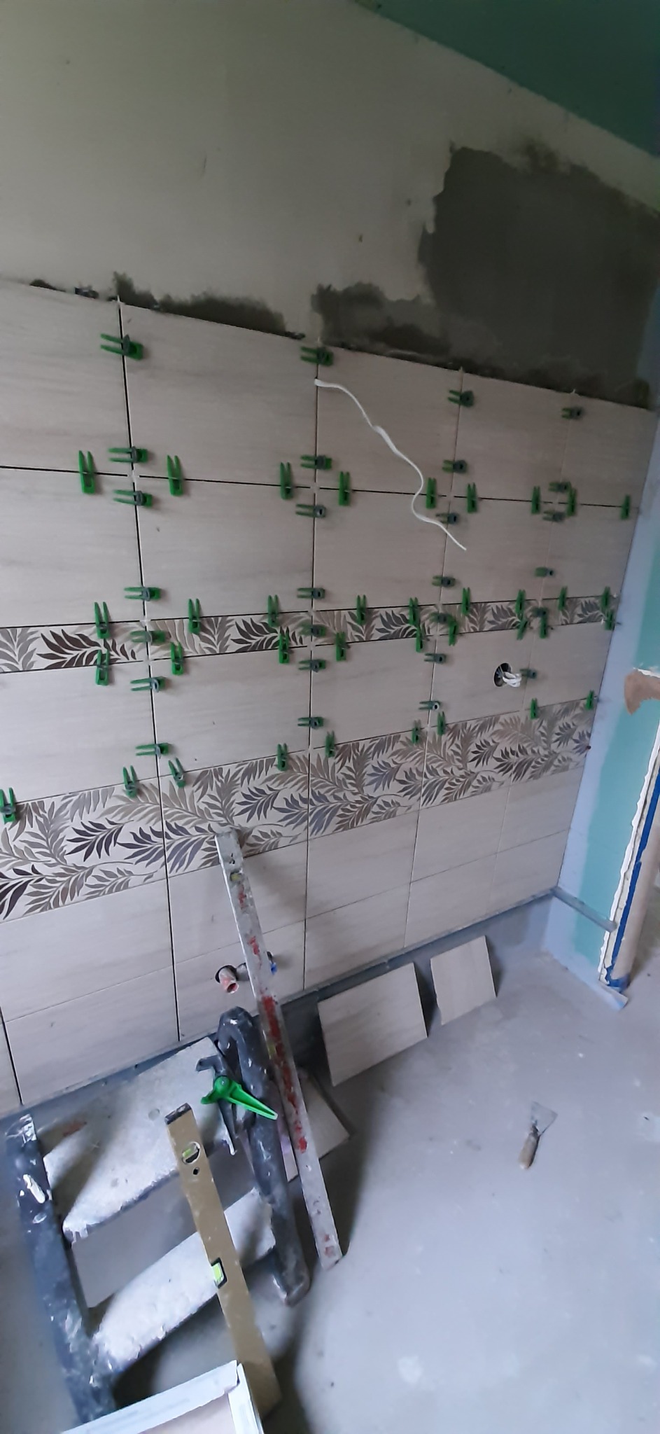 Układanie beżowych płytek z motywem liści na ścianie, z użyciem zielonych krzyżyków dystansowych i poziomicą opartą o ścianę, widoczne przewody elektryczne i fragment ściany z płyt...