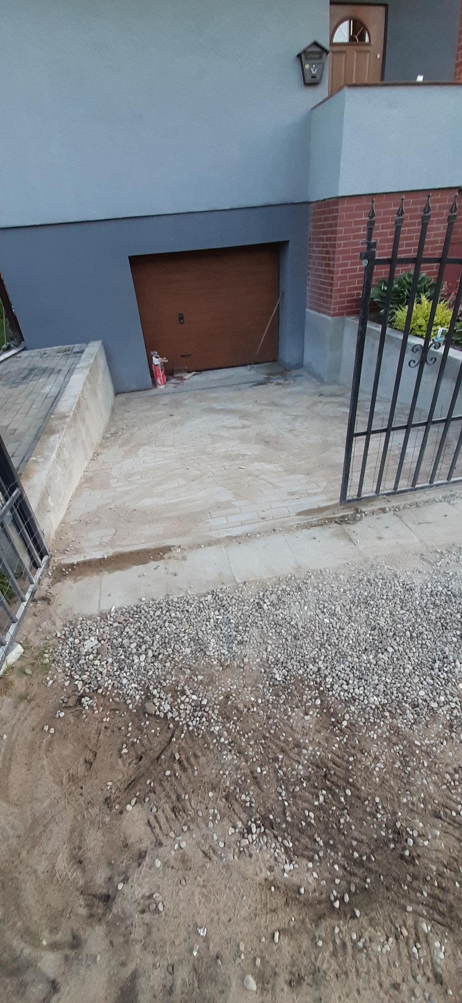 Ułożona kostka brukowa na podjeździe przed garażem, widoczne ślady po pracy, rozsypany żwir i resztki materiałów budowlanych.