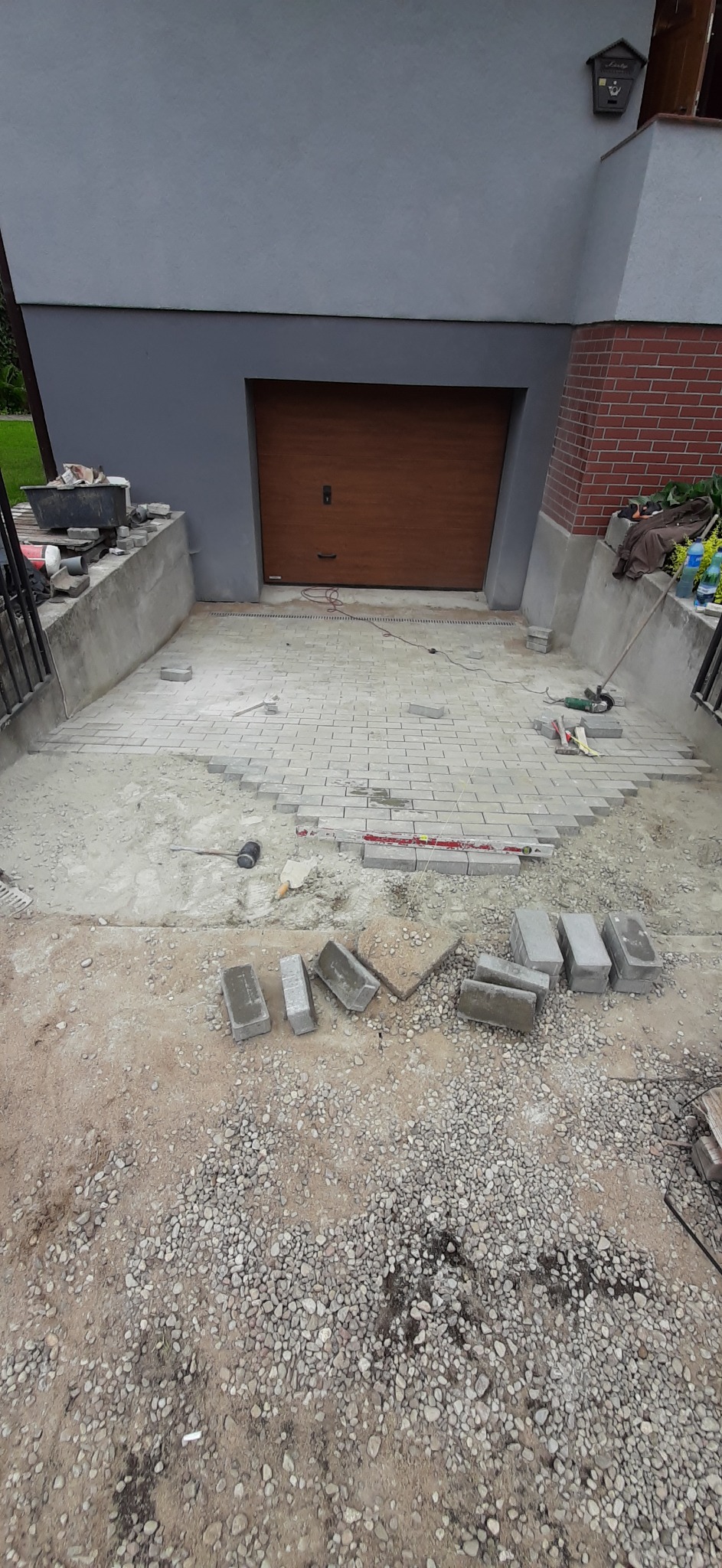 Układanie szarej kostki brukowej na podjeździe przed garażem, widoczne narzędzia i niedokończony fragment nawierzchni.
