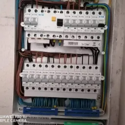 Otwarta skrzynka elektryczna z zainstalowanymi bezpiecznikami Eaton, widoczne okablowanie w kolorach brązowym, niebieskim, zielono-żółtym, z bliska na tle ściany.