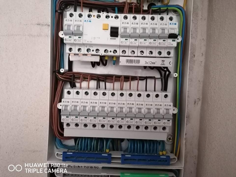 Otwarta skrzynka elektryczna z zainstalowanymi bezpiecznikami Eaton, widoczne okablowanie w kolorach brązowym, niebieskim, zielono-żółtym, z bliska na tle ściany.