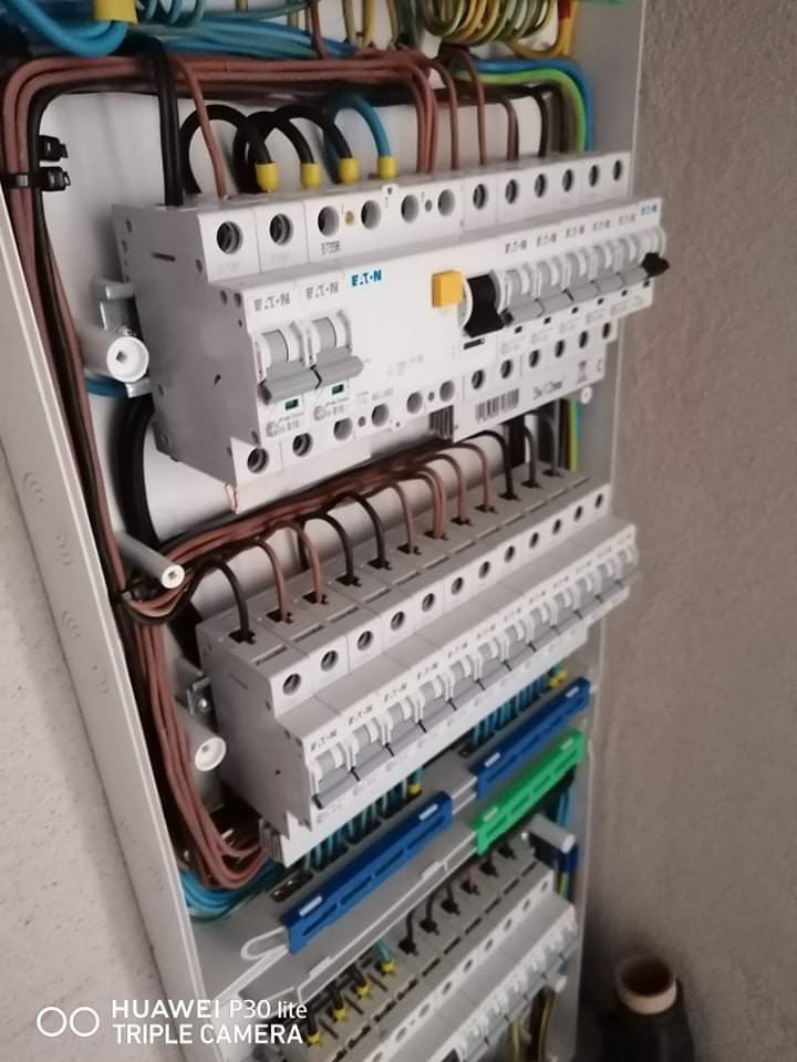 Otwarta skrzynka elektryczna z zainstalowanymi bezpiecznikami Eaton, widoczne kable w różnych kolorach i oznaczeniach, szyny DIN z niebieskimi i zielonymi elementami mocującymi.