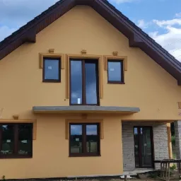 Żółta elewacja domu jednorodzinnego z brązowymi oknami i dachem, widoczne dekoracyjne elementy elewacyjne oraz słupy energetyczne w tle.