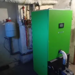 Nowoczesny, zielony piec CO w kotłowni. Widoczny zbiornik wyrównawczy i instalacja grzewcza. Ujęcie w piwnicy, oświetlenie sufitowe.