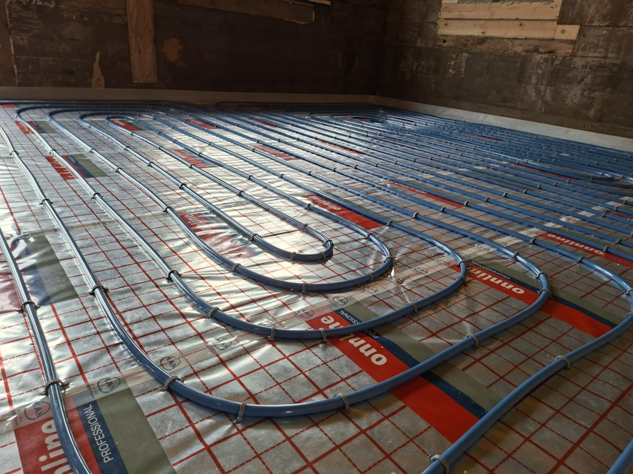 Niebieskie rury ogrzewania podłogowego ułożone na folii z czerwoną kratką, przymocowane plastikowymi uchwytami, widok z góry, surowy stan wykończenia.