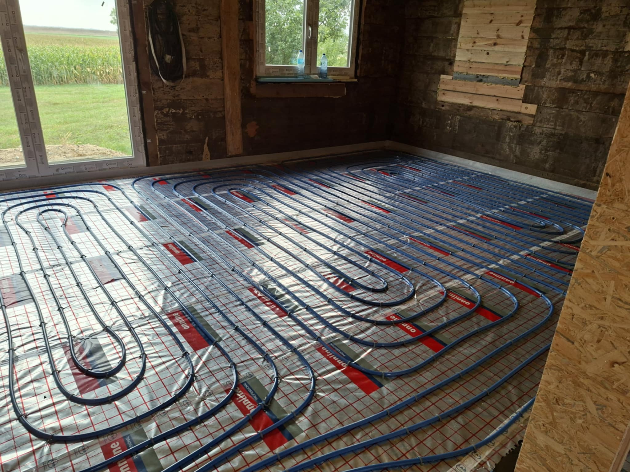 Instalacja ogrzewania podłogowego z niebieskich rurek na folii w surowym wnętrzu z oknem i widokiem na pole. Widoczne fragmenty ścian przed wykończeniem.