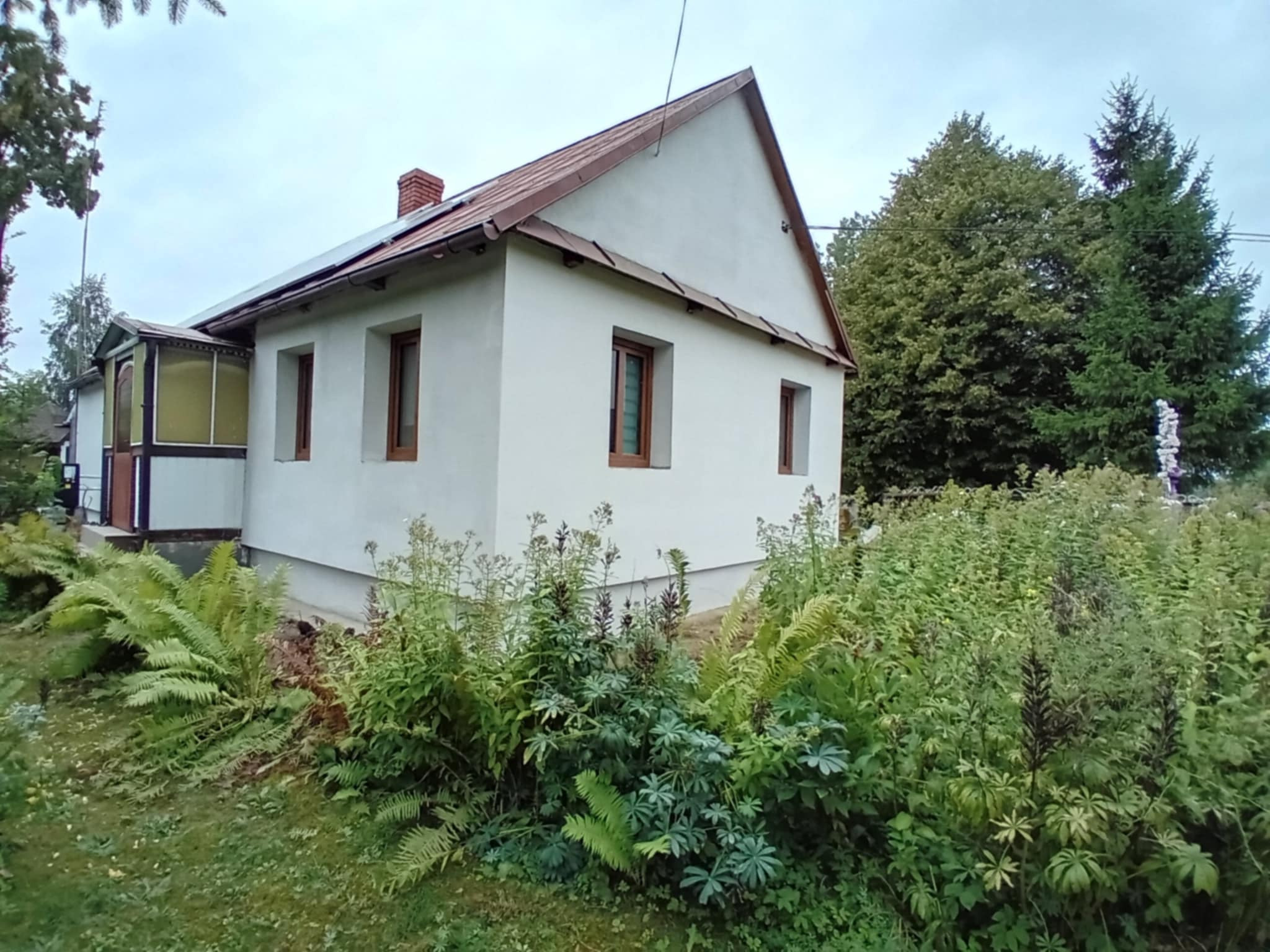Biały dom z brązowymi oknami i dachem, otoczony zielenią. Widoczna dobudówka z przeszkleniami i komin. Ujęcie z poziomu gruntu, zieleń na pierwszym planie.