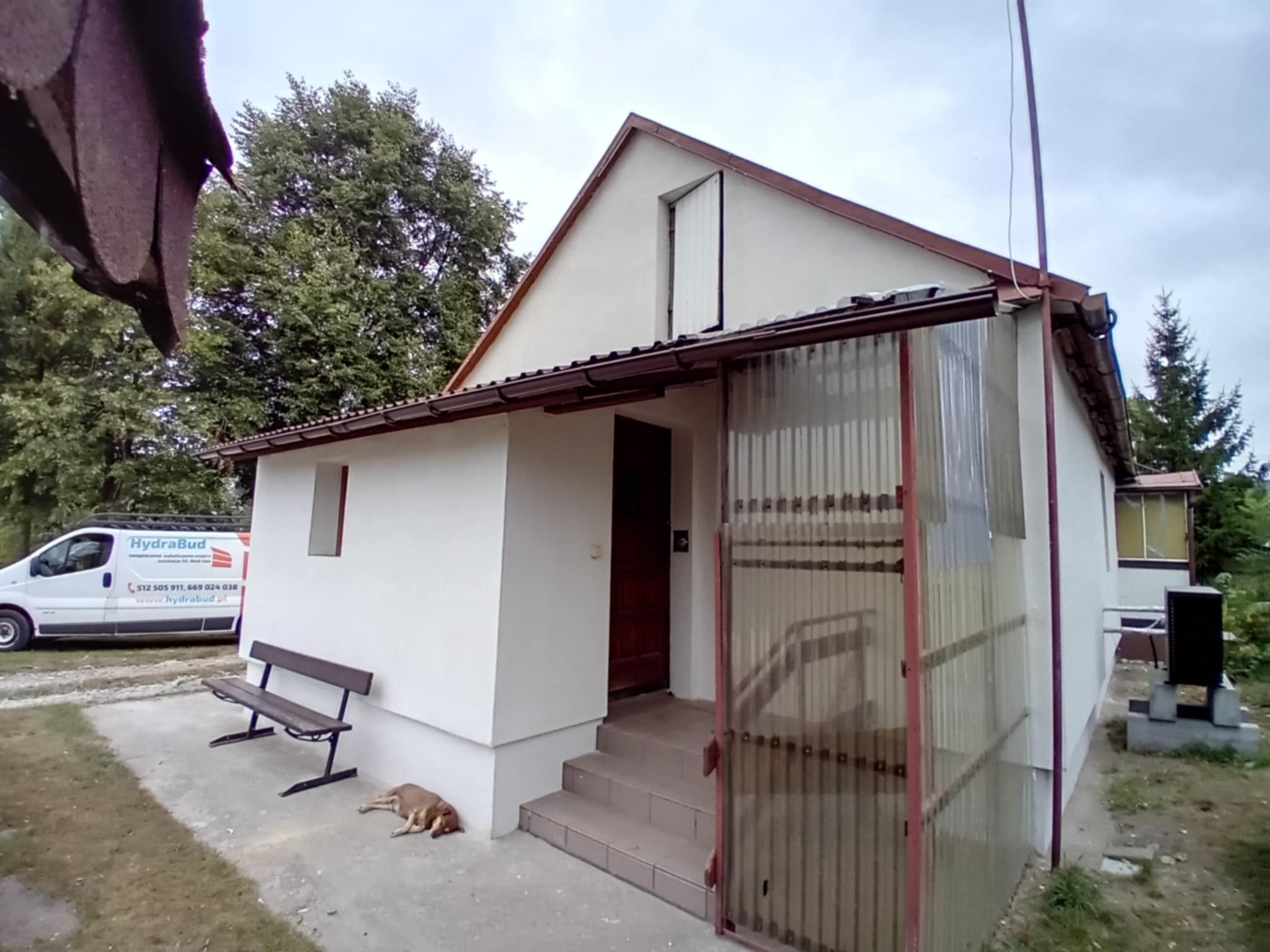 Dom jednorodzinny z jasną elewacją, widoczny z zewnątrz, z psem leżącym obok ławki. Na ścianie widoczny pojazd z logo firmy.