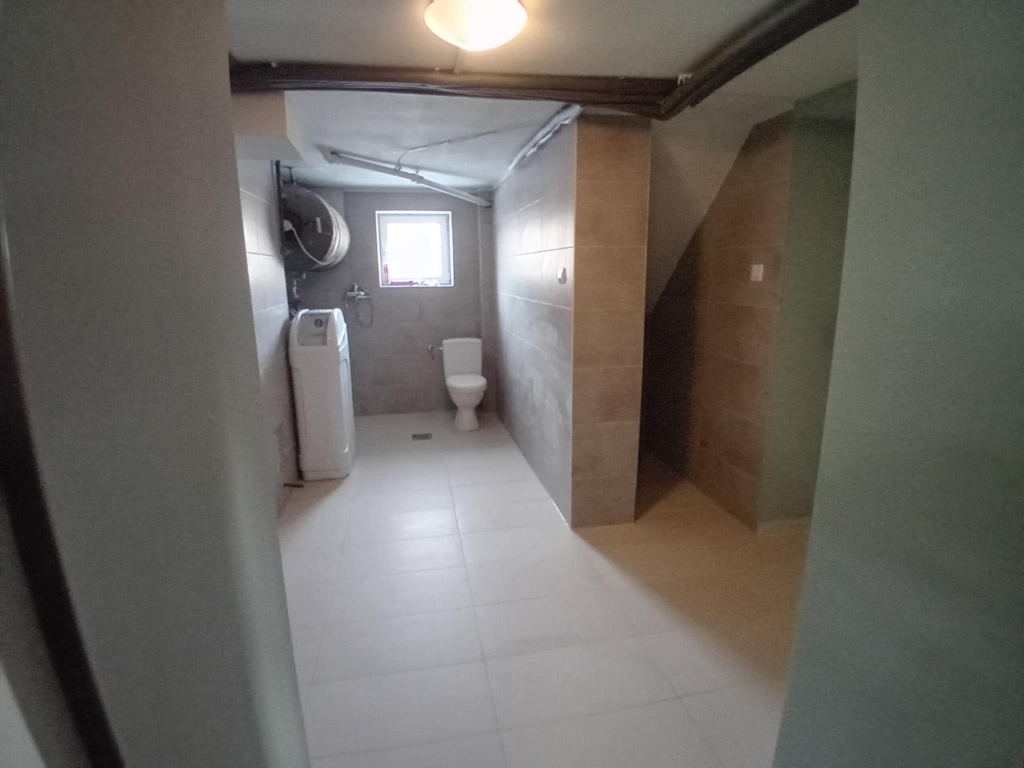 Wykończona łazienka w jasnych płytkach z toaletą, pralką i bojlerem. Widok z korytarza, oświetlenie sufitowe. Minimalistyczny design, świeżo po remoncie.