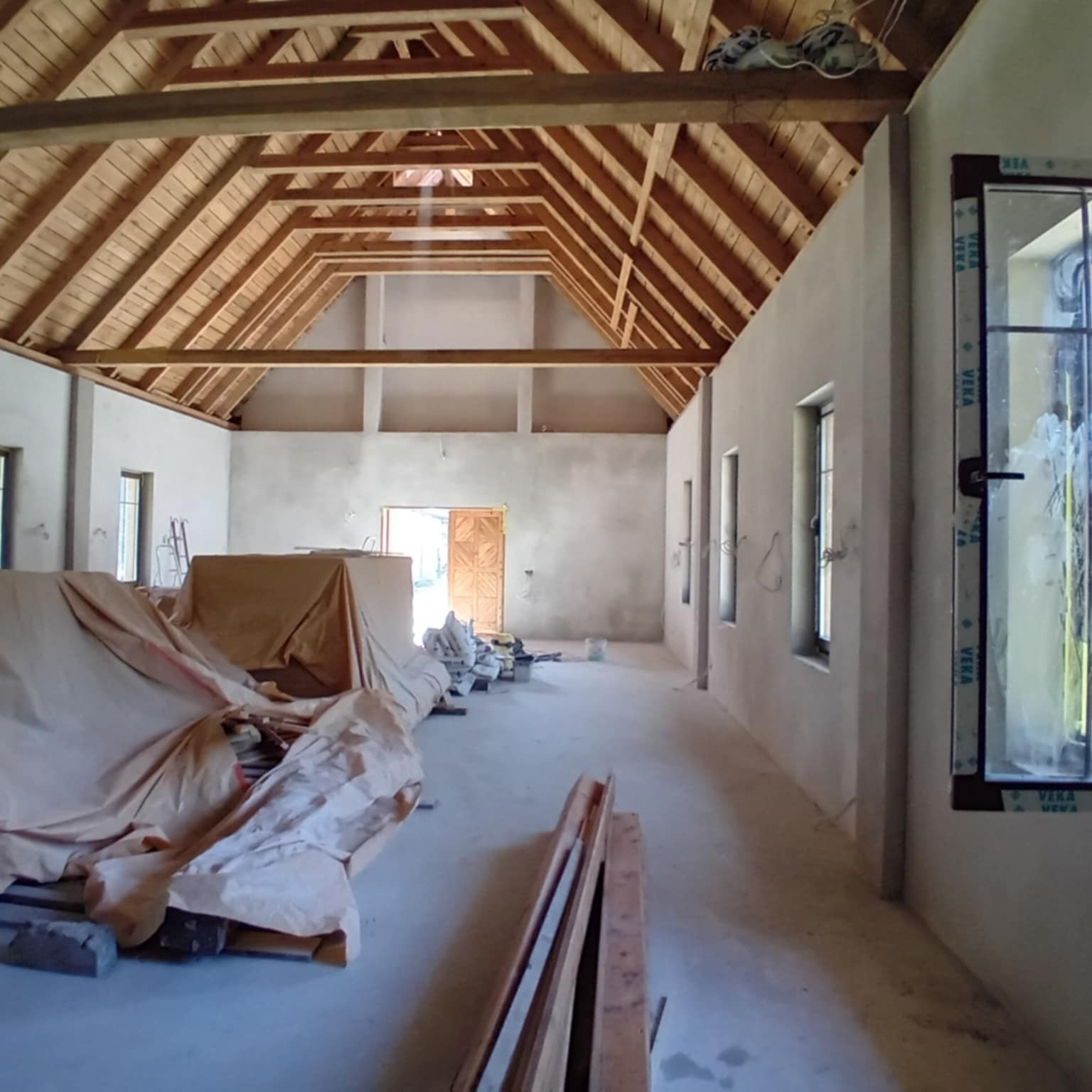 Wnętrze w trakcie prac wykończeniowych: surowy drewniany strop, ściany przygotowane pod malowanie, okna PCV z widocznymi zabrudzeniami, materiały budowlane pod przykryciem.