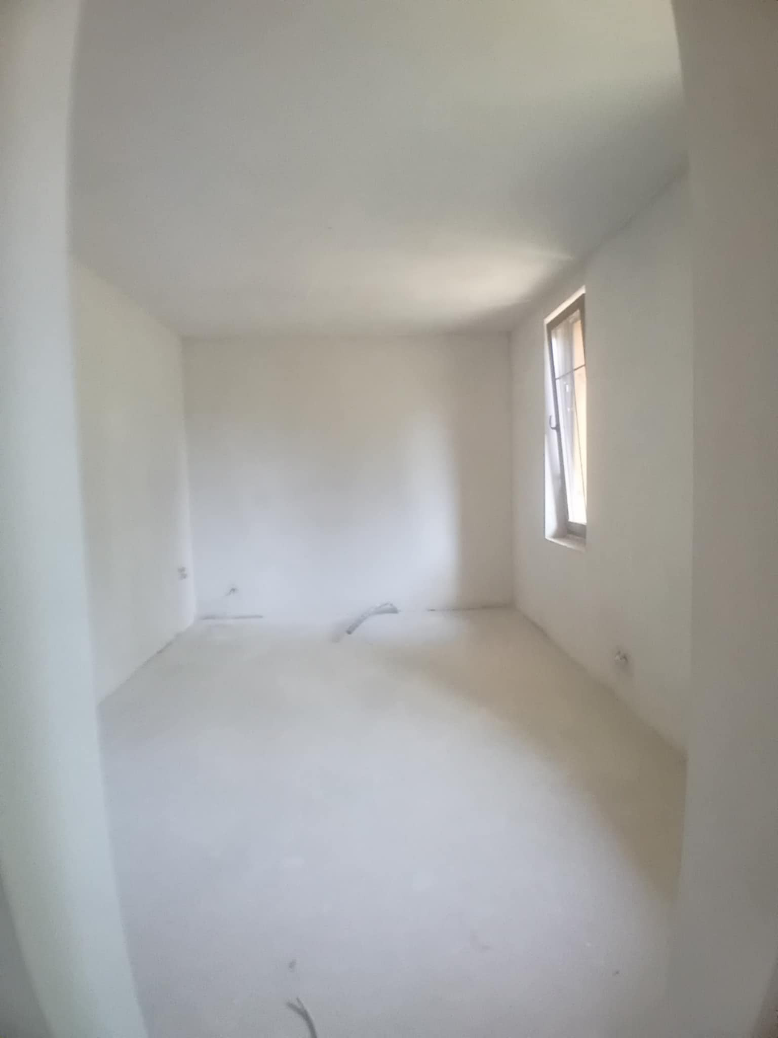 Puste, jasne pomieszczenie po szpachlowaniu i malowaniu na biało. Widoczne ślady po instalacjach na ścianach i podłodze. Okno w prawej ścianie.