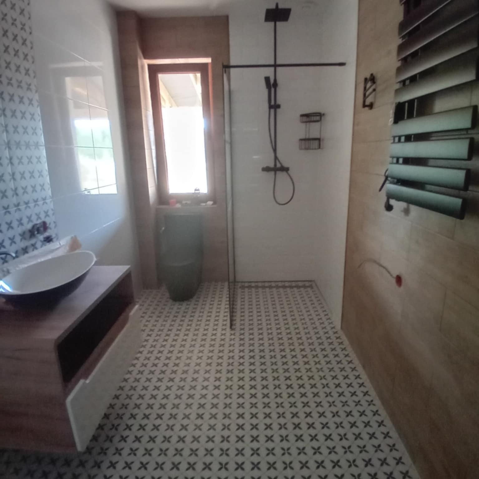 Nowoczesna łazienka z kabiną prysznicową typu walk-in, umywalką nablatową i wzorzystą podłogą. Widoczne okno z doniczką i grzejnik drabinkowy.