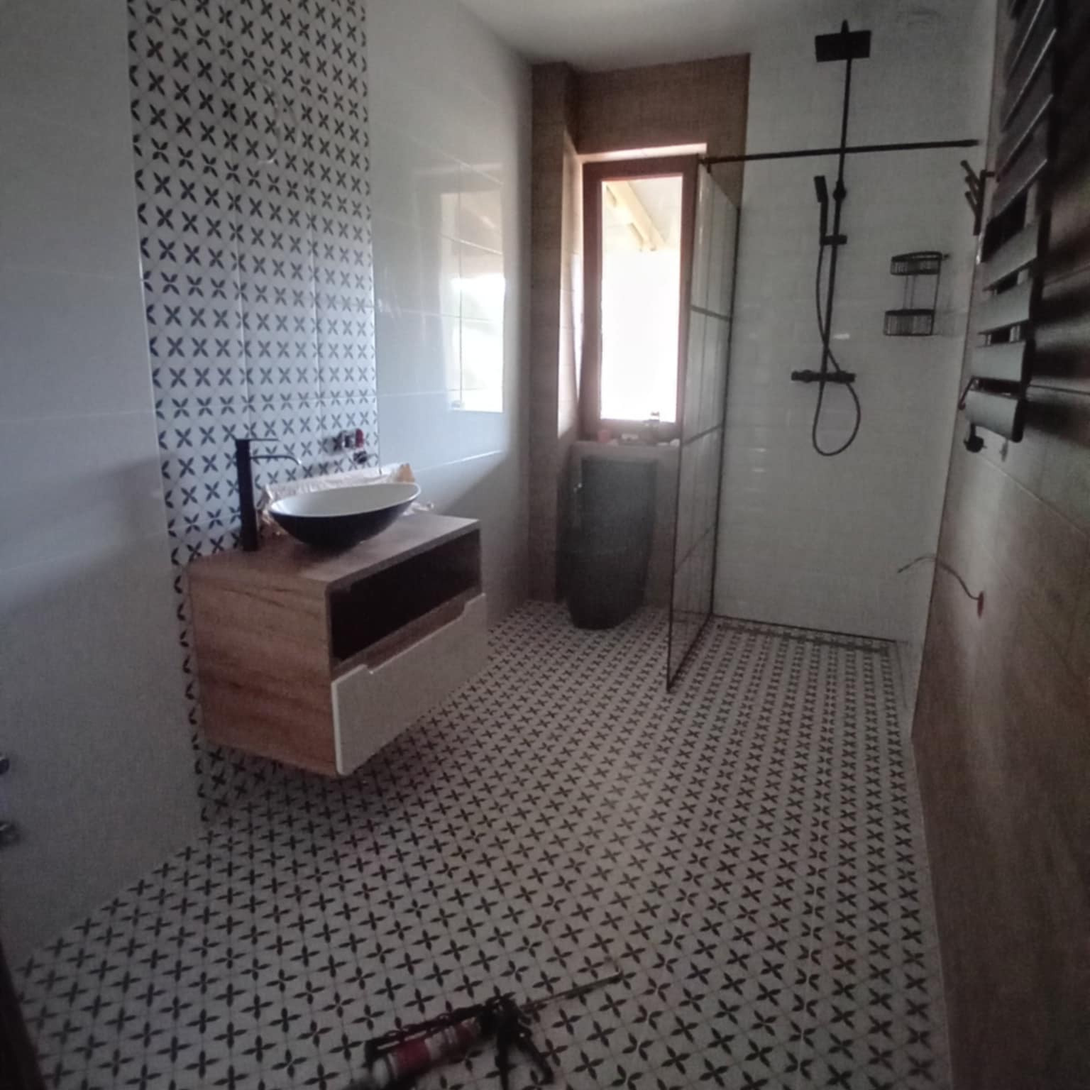 Nowoczesna łazienka po remoncie: białe płytki ze wzorem krzyżyków na podłodze i ścianie, drewniana szafka z umywalką, czarna armatura, kabina prysznicowa.