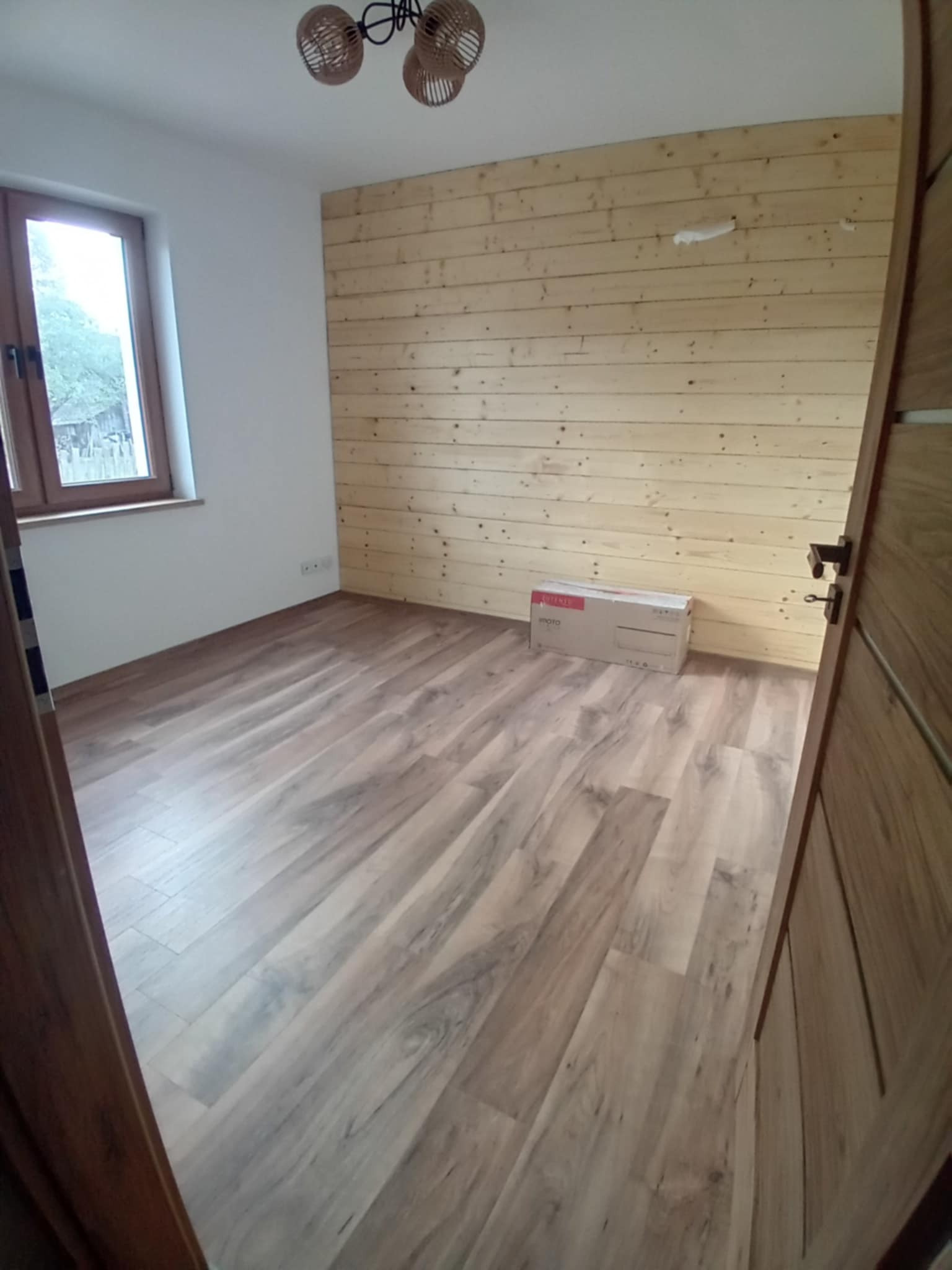 Pokój po remoncie: drewniana ściana, panele podłogowe imitujące drewno, okno z widokiem na zewnątrz, lampa wisząca. W rogu pudełko.