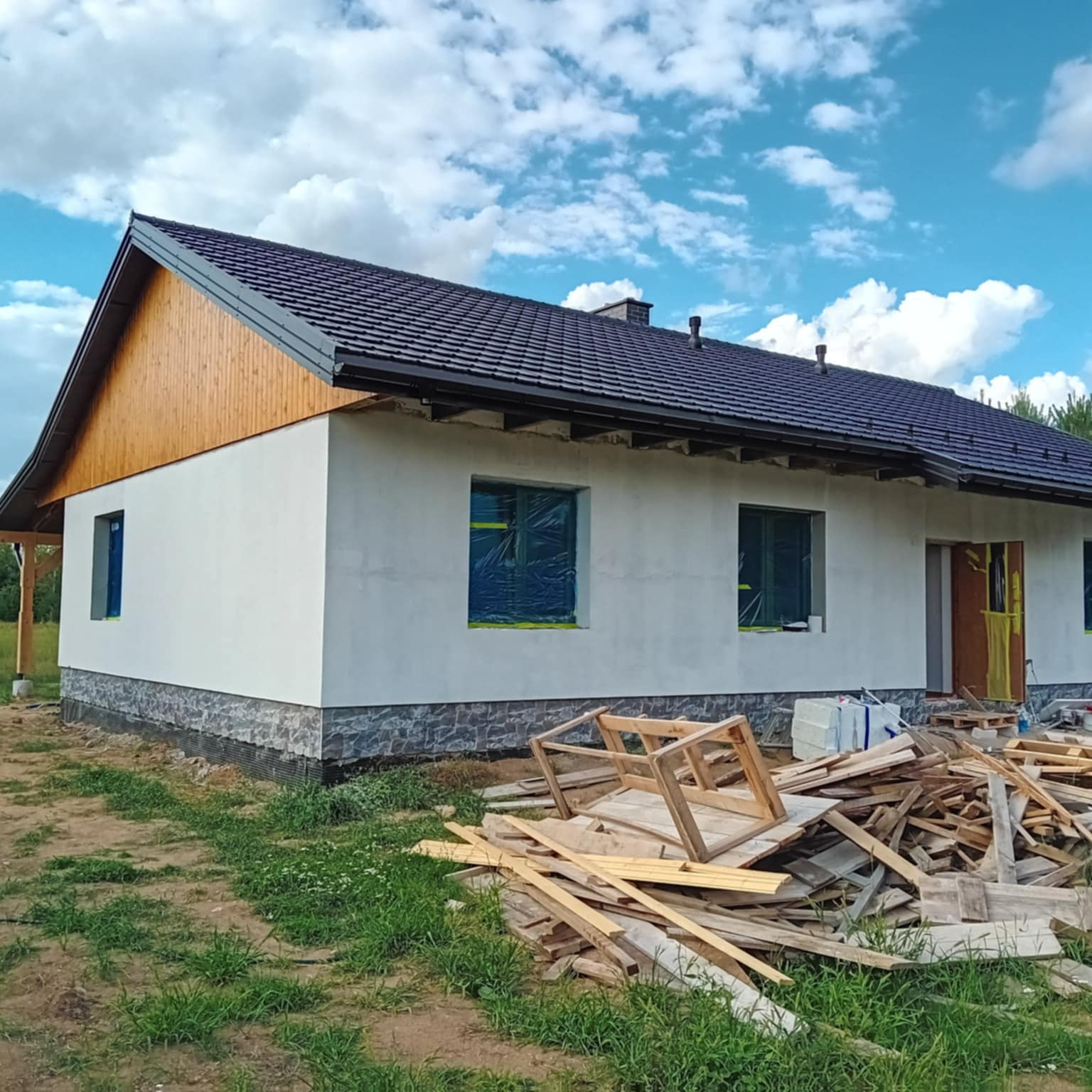 Parterowy dom z białą elewacją, ciemnym dachem i drewnianymi elementami wykończeniowymi. Okna zabezpieczone folią. Na pierwszym planie stos drewna.