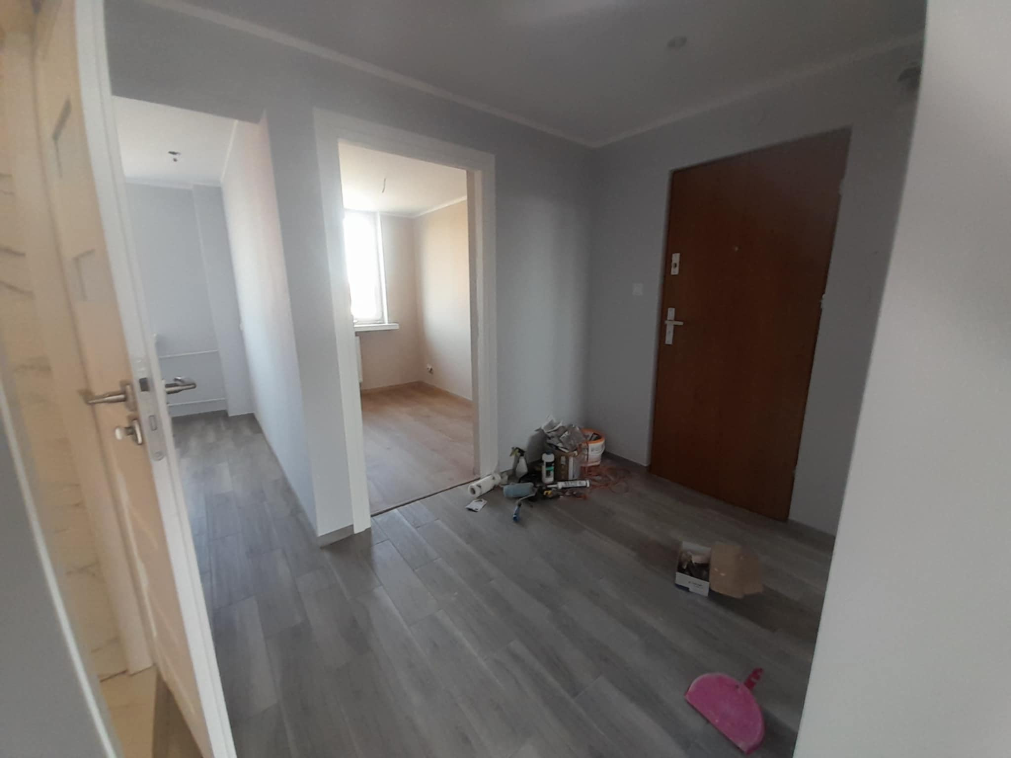 Wnętrze po remoncie w Hajnówce: szare panele podłogowe, pomalowane ściany na szaro, widoczne narzędzia malarskie. Jasne pomieszczenie, drzwi wejściowe.