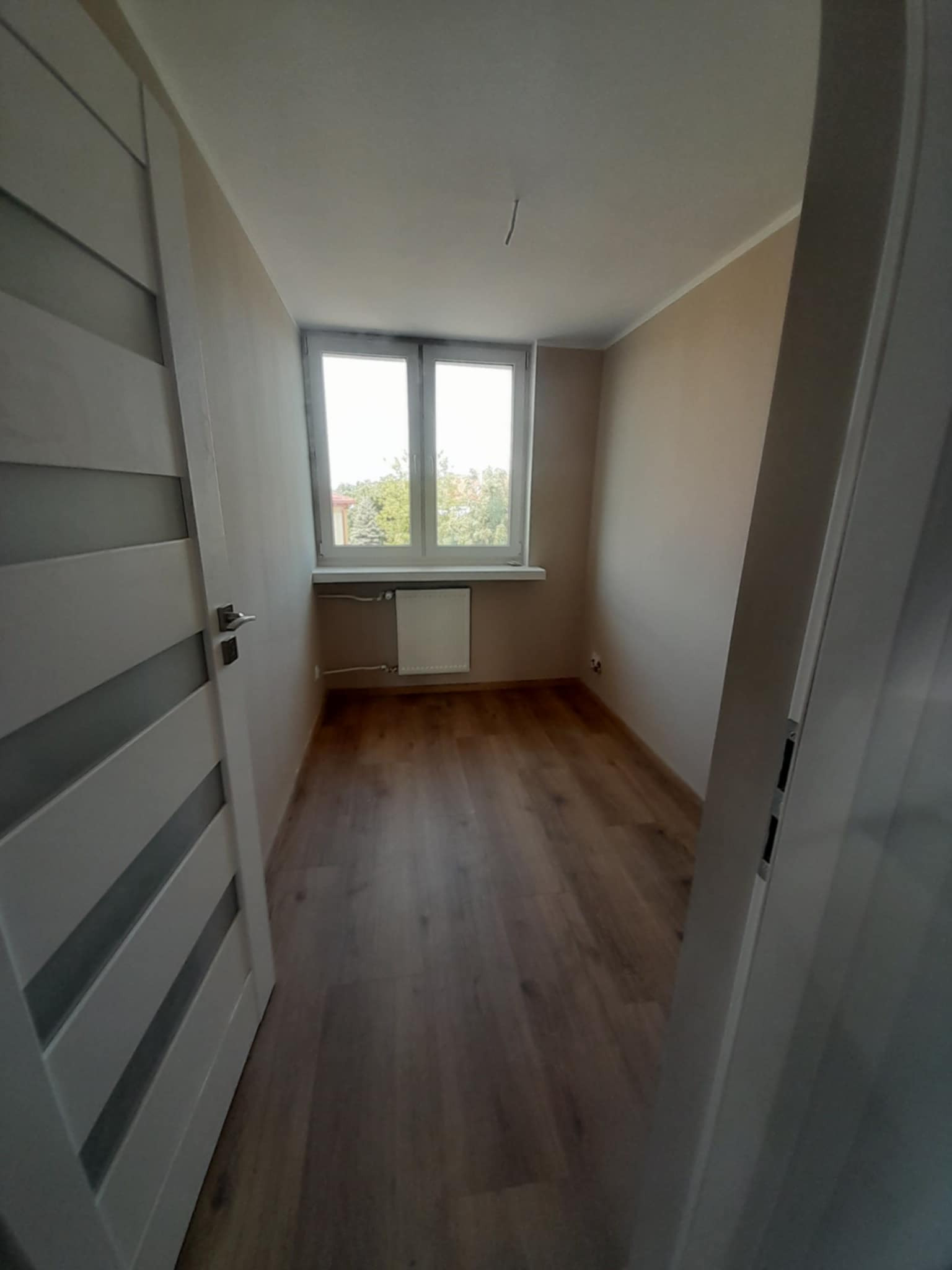 Wykończone wnętrze pokoju z drewnianą podłogą i świeżo pomalowanymi ścianami w odcieniach beżu, widoczne drzwi z przeszkleniami i okno z widokiem na zieleń.