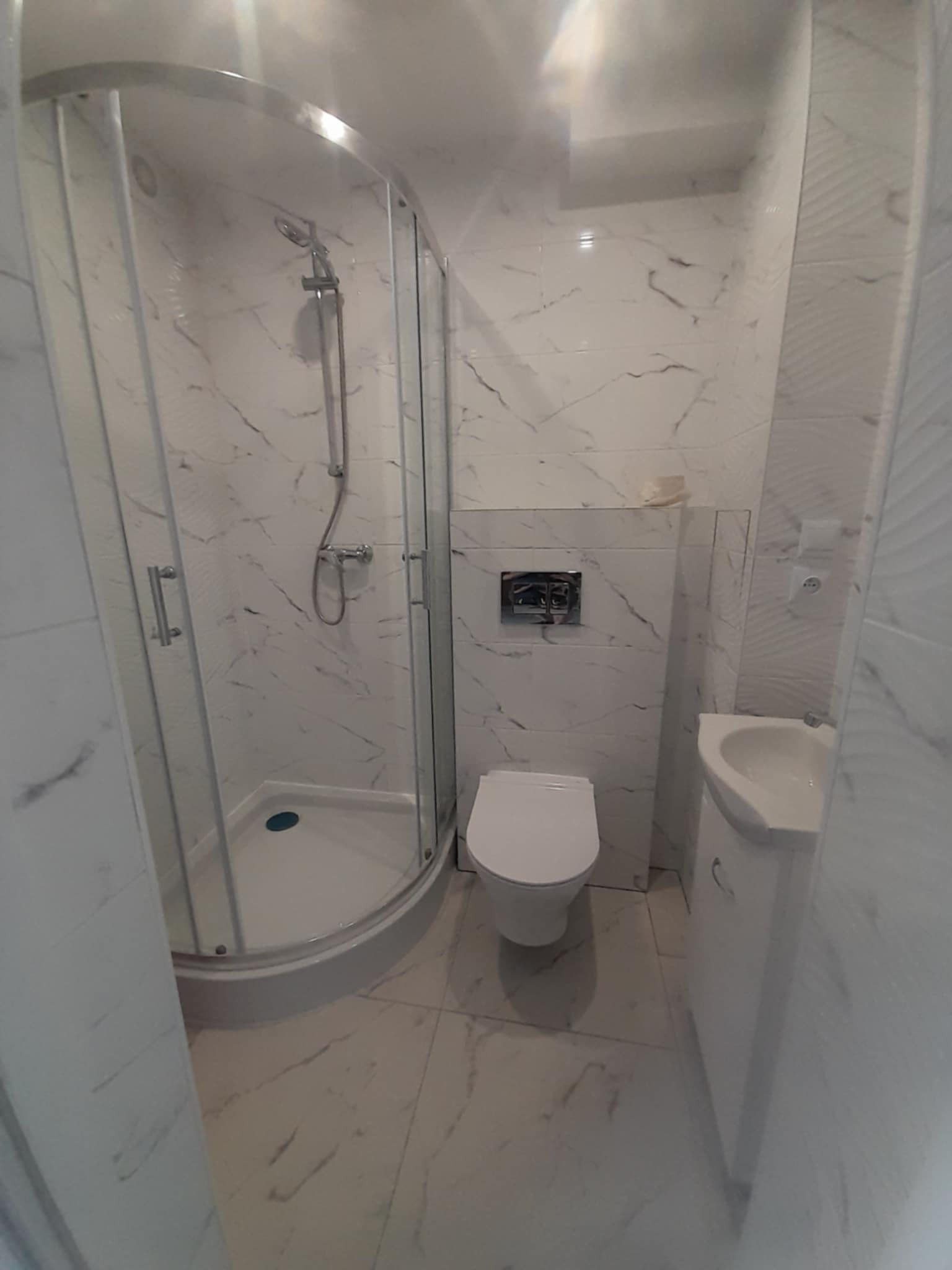 Wykończona łazienka w bieli: kabina prysznicowa z chromowaną armaturą, podwieszana toaleta i umywalka z szafką, całość w płytkach imitujących marmur.