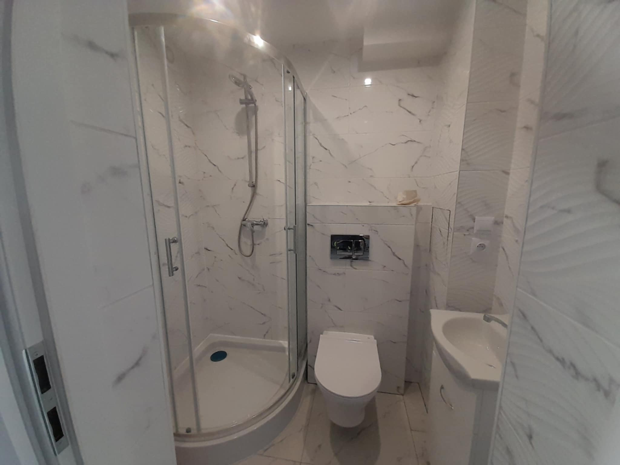 Wykończona łazienka w bieli z kabiną prysznicową, umywalką i toaletą. Ściany i podłoga wyłożone płytkami imitującymi marmur. Nowoczesny design, minimalistyczny styl.