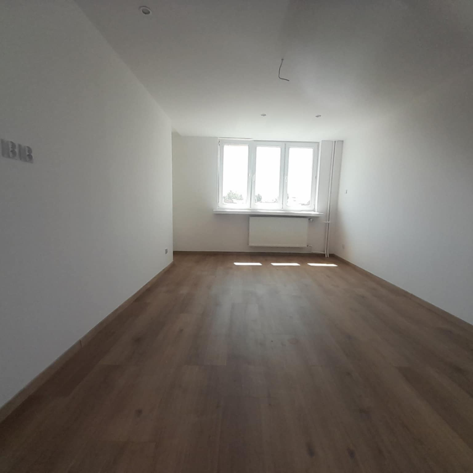 Puste, jasne pomieszczenie po remoncie z drewnianą podłogą i białymi ścianami. Widoczne gniazdka elektryczne, okno z grzejnikiem i sufit z oświetleniem.