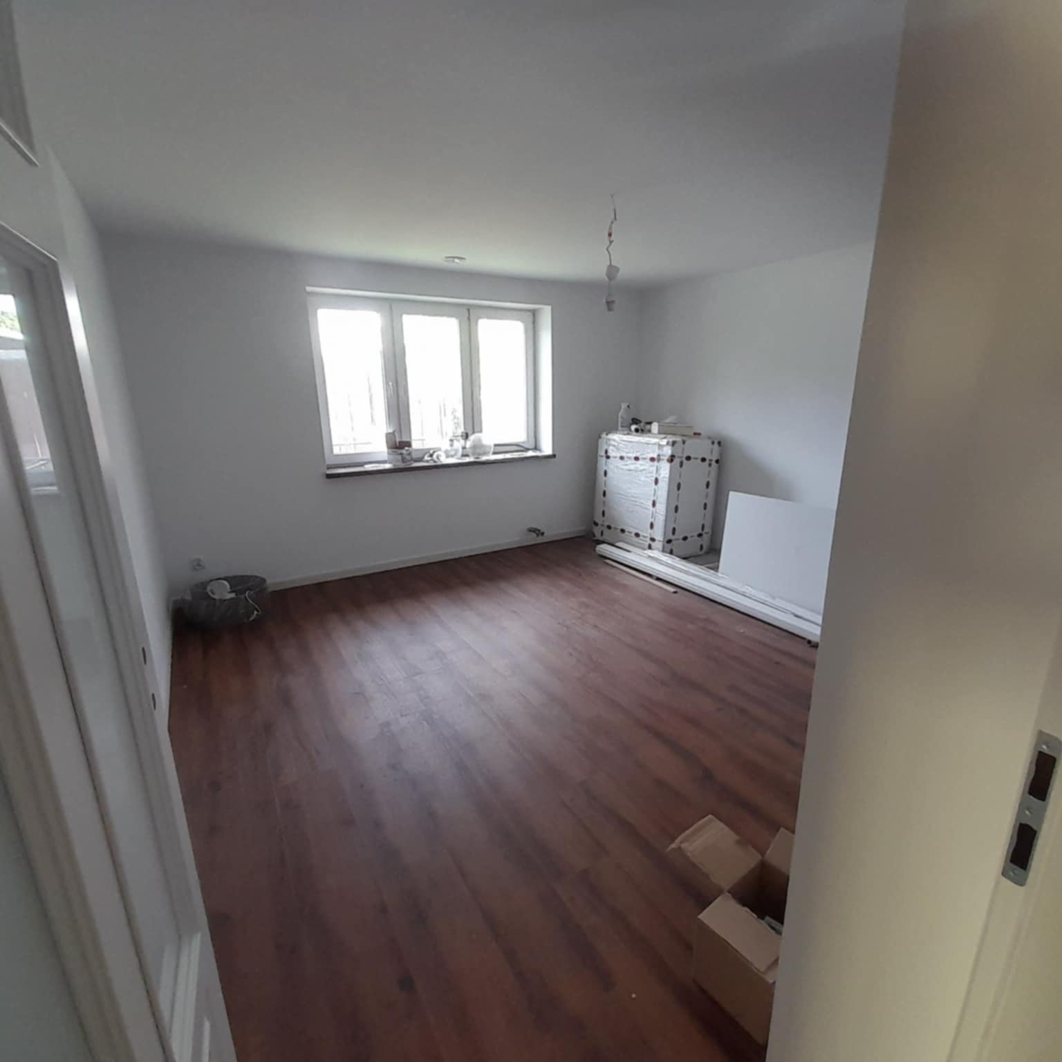 Pokój po remoncie z drewnianą podłogą, białymi ścianami i oknem. Widoczne materiały budowlane i karton. Ujęcie z perspektywy otwartych drzwi.