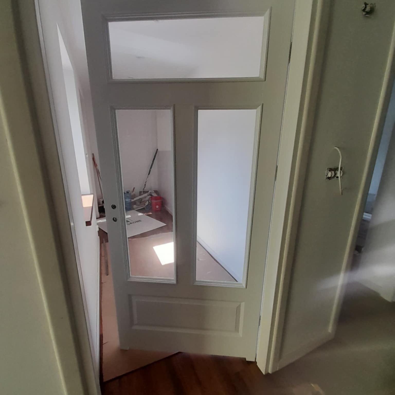 Otwarta, biała, przeszklona wewnętrzna stolarka drzwiowa. Widoczne zabezpieczone podłoże i narzędzia w remonowanym pomieszczeniu. Ujęcie z perspektywy podłogi.