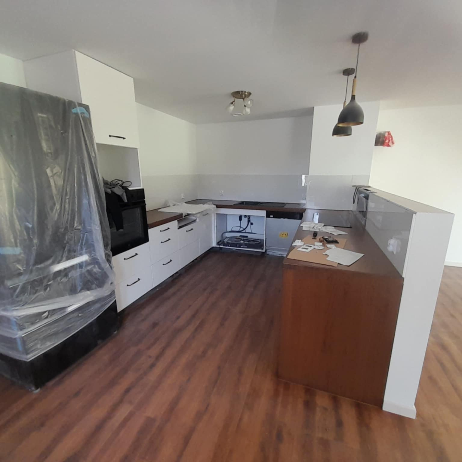 Kuchnia w trakcie remontu: białe szafki, drewniany blat, podłoga imitująca drewno, sprzęt AGD zabezpieczony folią, przygotowania do montażu.