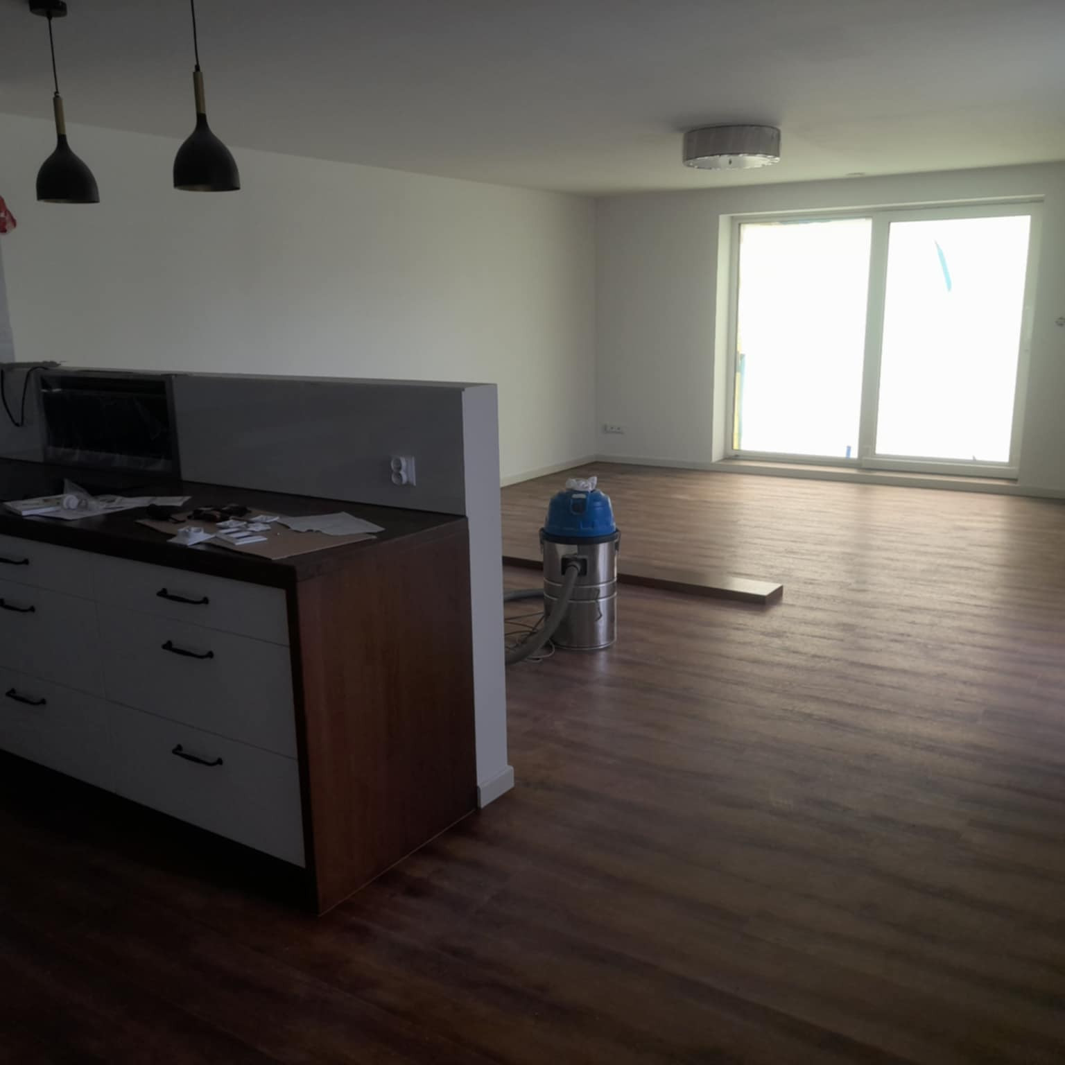 Wnętrze pokoju z ułożoną podłogą z paneli o ciemnym odcieniu drewna. Widoczny odkurzacz przemysłowy i fragment zabudowy kuchennej. Duże okno z dostępem do światła dziennego.
