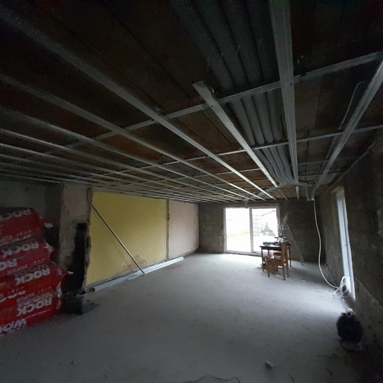 Wnętrze w trakcie prac wykończeniowych: metalowa konstrukcja sufitu podwieszanego, izolacja ROCKWOOL, widoczne rury wentylacyjne pod stropem. Stan surowy.