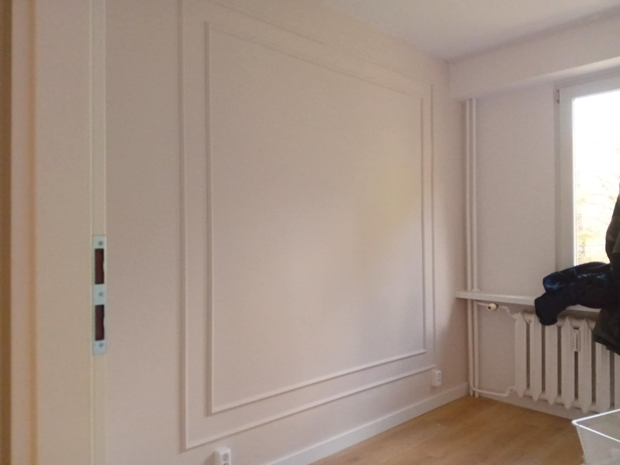 Jasny pokój po remoncie: ściana z dekoracyjnymi listwami w ramce, drewniana podłoga, grzejnik, okno. Minimalistyczny design, subtelne detale wykończenia.