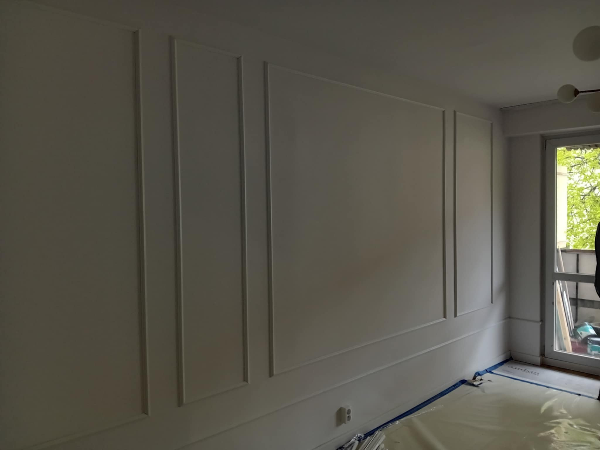 Biała ściana z dekoracyjnymi listwami w pionowe prostokąty, wykończona sztukateria, osłonięta folią malarską. Widok na okno z zielenią za nim.