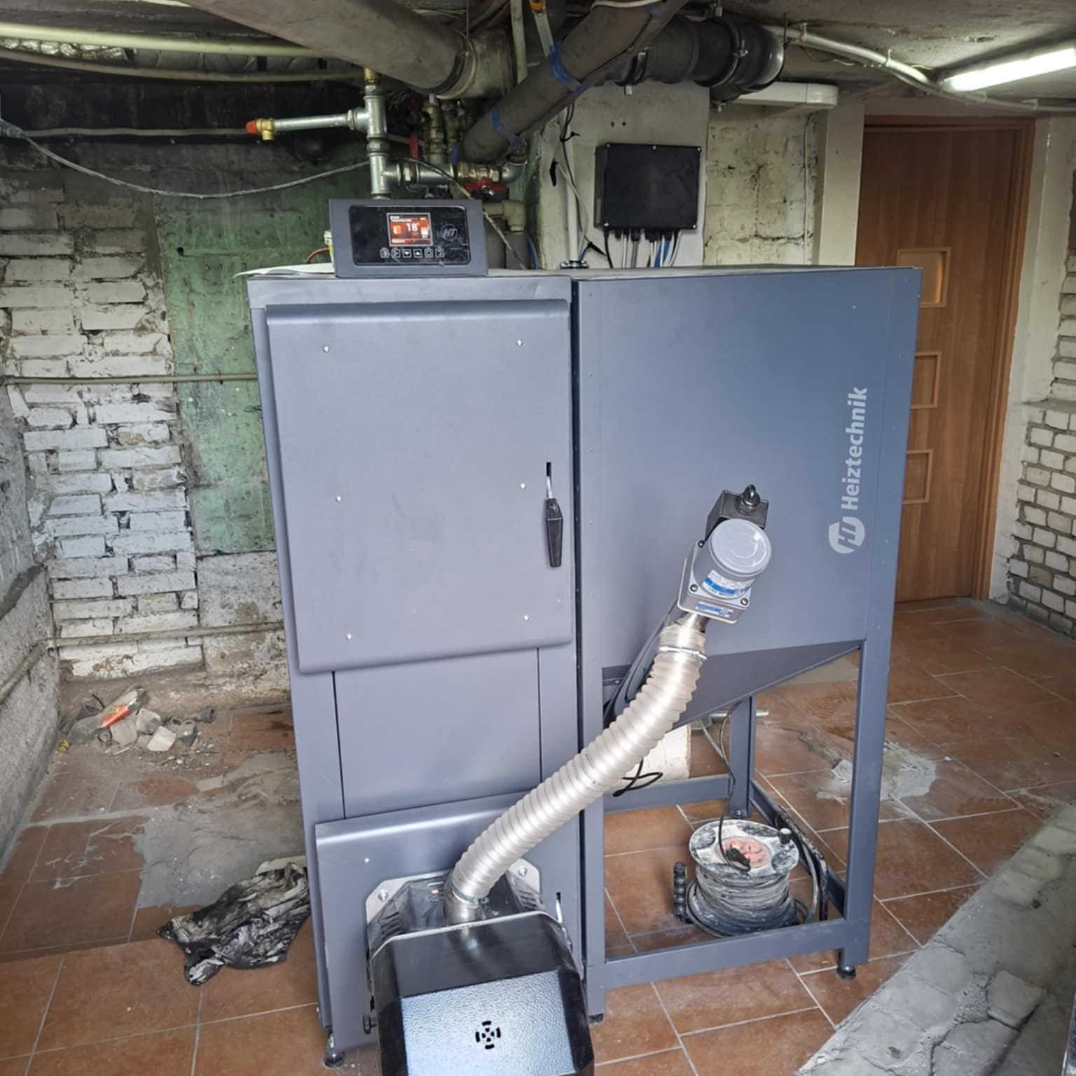Szary piec Heitztechnik w piwnicy z surowymi ścianami i instalacją pod sufitem. Widoczny panel sterowania i podłączenia. Na podłodze płytki i resztki gruzu.