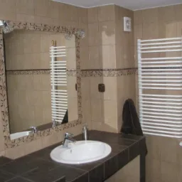 Beżowa łazienka z ciemnobrązowym blatem, umywalką, lustrem z motywem roślinnym i białym grzejnikiem drabinkowym.