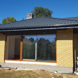 P.H.U. Okneks Łukasz Kamiński - Montaż dużych okien z drewnianymi ramami w nowo budowanym domu z żółtej cegły, widoczne rusztowanie i przygotowane materiały budowlane.