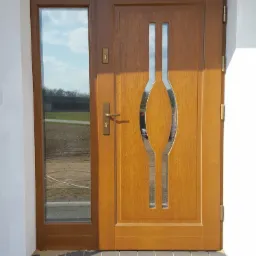 Brązowe drzwi wejściowe z bocznym przeszkleniem i ozdobnymi, pionowymi intarsjami ze stali nierdzewnej, zamontowane w jasnym budynku.