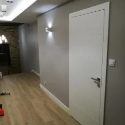 Nowoczesne, białe drzwi wewnętrzne z poziomymi frezowaniami, zamontowane w szarej ścianie, widok na korytarz z oświetleniem punktowym i podłogą imitującą drewno.