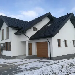 Nowoczesny dom jednorodzinny z antracytowym dachem, jasną elewacją i drewnianą bramą garażową w zimowej scenerii z resztkami śniegu na podjeździe.