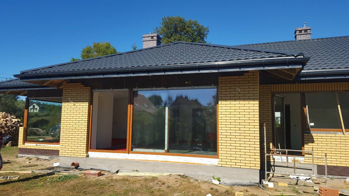 Montaż dużych okien z drewnianymi ramami w nowo budowanym domu z żółtej cegły, widoczne rusztowanie i przygotowane materiały budowlane.