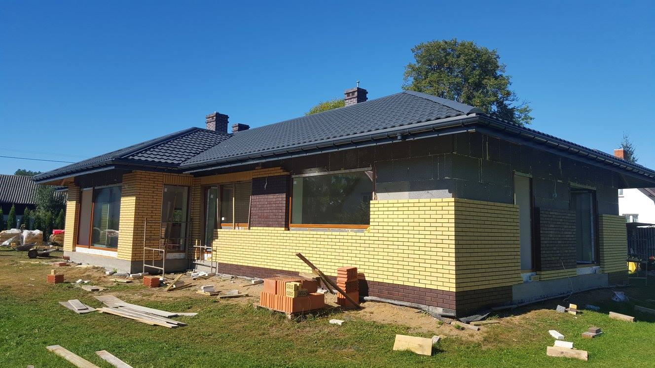 Dom jednorodzinny w trakcie budowy, widoczne okna z drewnianą ramą, fragmenty ocieplenia i murowana elewacja z żółtej cegły klinkierowej.
